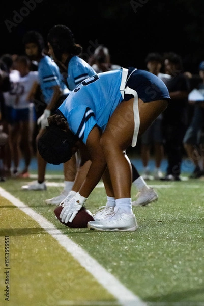 Fototapeta Female flag football center prepares to snap under the stadium lights. Focused stance, ready hands, and team alignment showcase precision and preparation in youth sports.