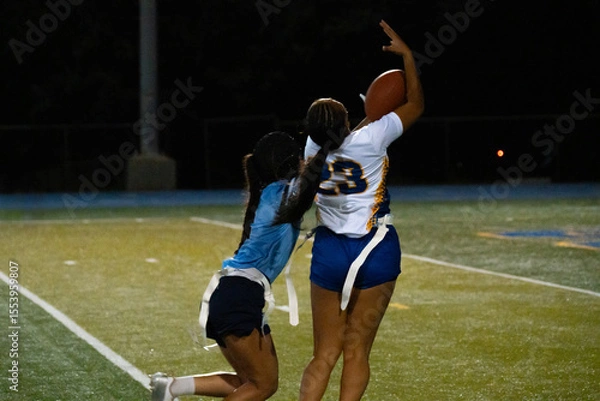 Fototapeta Female flag football receiver leaps for catch under pressure, with defender closing in during intense nighttime match. Athleticism, focus, and sportsmanship captured on artificial turf