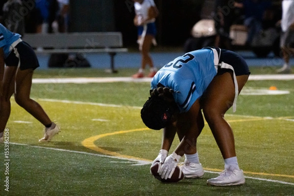 Fototapeta Girls flag football center prepares to snap the ball under lights, showing discipline, focus, and readiness in youth sports competition. Team positioning and intensity captured on turf at night.