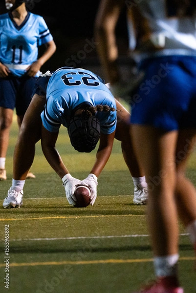 Fototapeta Girls flag football center prepares to snap the ball at night game, crouched in position with focus and intent, youth athletes in action under lights, sports readiness, school competition scene.