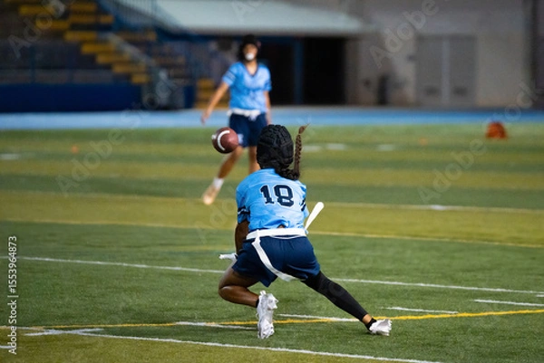 Fototapeta Girls flag football receiver in blue jersey crouches to catch pass during night game, intense sports action on turf, youth athletic focus, school competition, teamwork and coordination captured.