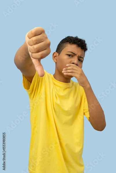 Fototapeta Vertical image of a dark skinned Latino teenager wearing a yellow shirt with a disappointed face making a thumbs down gesture on a light blue background with copyspace