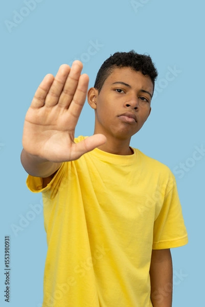 Fototapeta Vertical image of young Latino man wearing a yellow shirt with a serious face making a stop sign with his hand on a light blue background, teenager with a warning sign