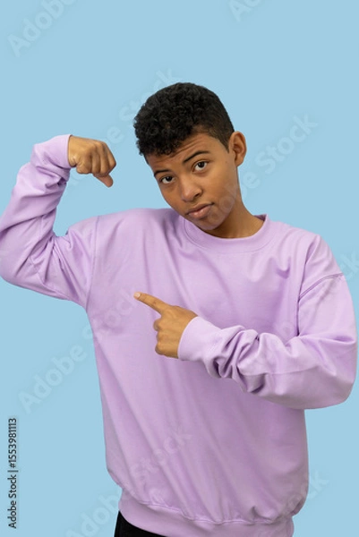 Fototapeta Vertical image of a young brown skinned Latino man making a gesture of strength with his hands wearing a lilac sweatshirt, pointing his muscles with his index finger on a light blue background