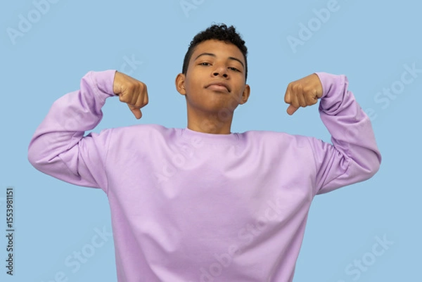 Fototapeta Young brown skinned Latino man making a gesture of strength with his hands, he is wearing a lilac sweatshirt on a light blue background, concept of youthful strength