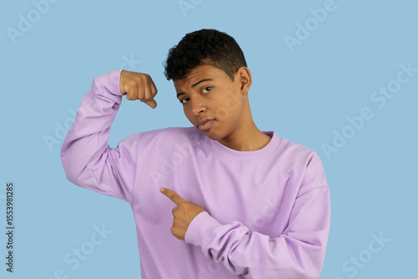 Fototapeta Young brown skinned Latino man making a gesture of strength with his hands, he is wearing a lilac sweatshirt and points his index finger at the muscle on a light blue background