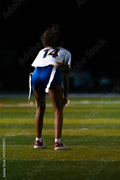 Fototapeta A female flag football player in ready stance under stadium lights. Number 14 on defense, focused and alert before the snap during a competitive night game.