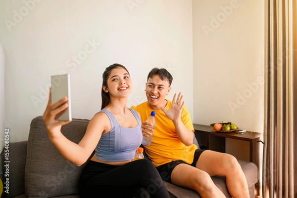 Fototapeta Indonesian southeast asian couple drinking water while taking a selfie or video call with their cellphone after finishing a home body workout exercise session. Fitness and active lifestyles concept