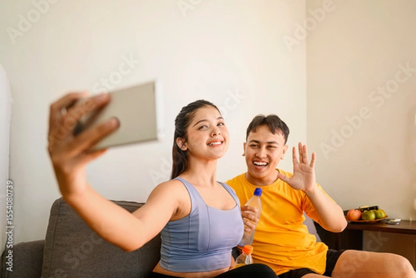 Fototapeta Indonesian southeast asian couple drinking water while taking a selfie or video call with their cellphone after finishing a home body workout exercise session. Fitness and active lifestyles concept