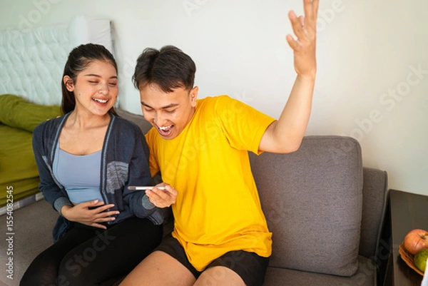 Fototapeta Indonesian southeast asian couple sitting on a sofa at home while looking results of a pregnancy test. Healthcare and family planning concept