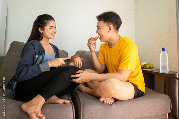 Fototapeta Indonesian southeast asian wife with a knife serves a slice of fruit to her husband while sitting on the sofa at home. Healthy eating concept