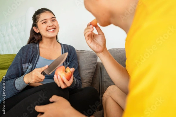 Fototapeta Indonesian southeast asian wife with a knife serves a slice of fruit to her husband while sitting on the sofa at home. Healthy eating concept