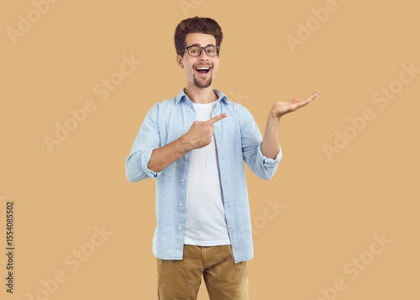 Fototapeta Laughing happy bearded man in glasses pointing index fingerand hand on copy space on beige background. Banner for advertisement, marketing, sales, offer, discount. Sincere human emotions concept.