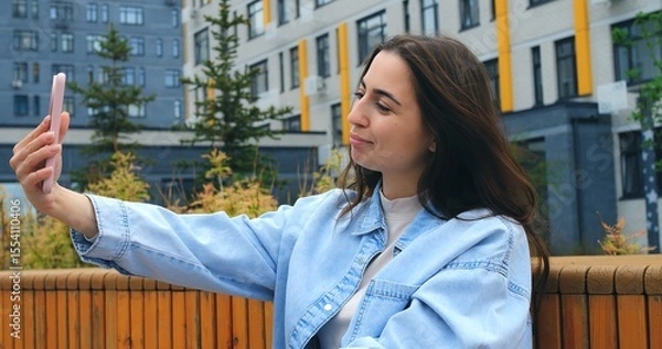 Obraz Outdoors, a young woman with long brown hair, wearing a light denim jacket, holds her smartphone and speaks into it, likely recording a voice message or using a voice assistant