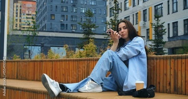 Obraz Young woman, wearing casual attire, sits on a wooden bench, savoring a takeaway coffee while engaging with her smartphone against a backdrop of modern buildings