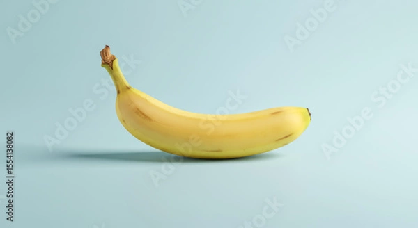 Fototapeta A single yellow banana lies on a light blue surface with soft lighting.