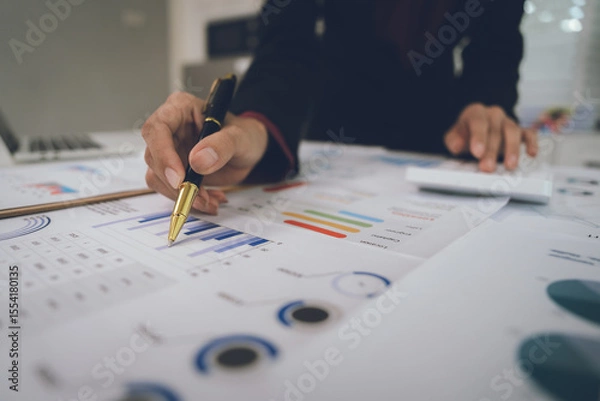 Fototapeta A businesswoman analyzes financial documents with charts and graphs at a desk, highlighting data trends with a pen in a bright, professional office setting.