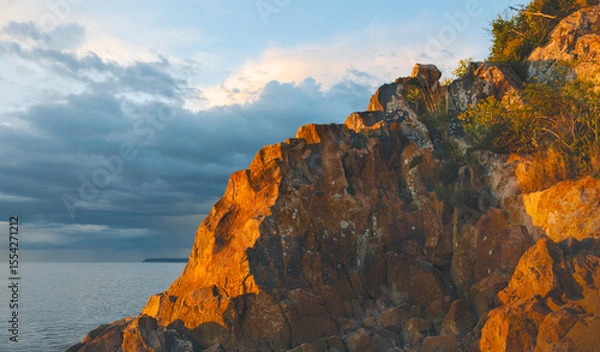 Fototapeta Rocky coastline glows under the evening light beside a calm sea and cloudy sky