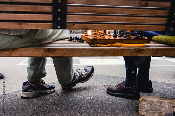 Fototapeta Salzburg in Austria. Two people sitting on a bench with a chess board in front of them. One of the people is wearing a pair of shoes