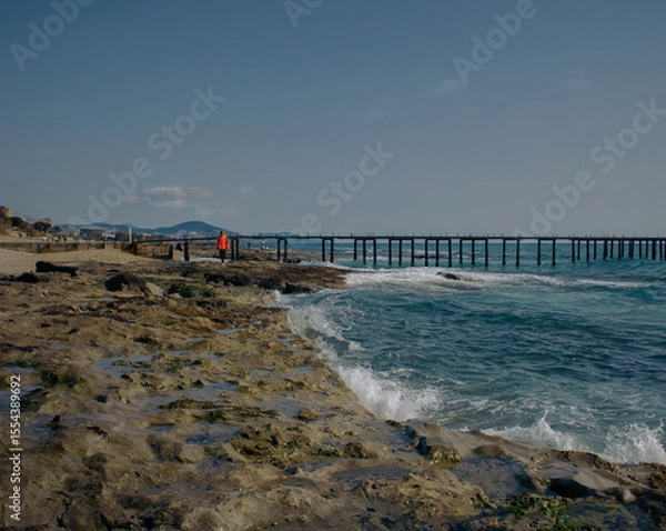 Fototapeta Winter sea with rocky beach and waves, figure in red coat
