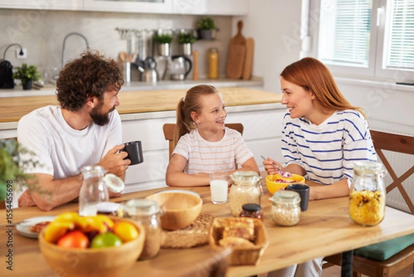Fototapeta A family of three sits at a wooden table in a modern kitchen, happily sharing breakfast on a weekend morning, filled with laughter and warm conversation over healthy meals.