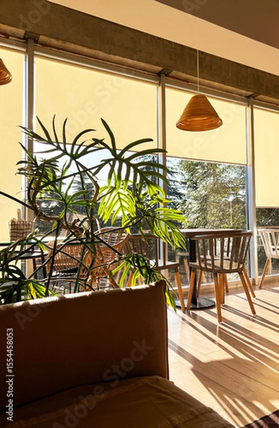 Fototapeta Bright and airy interior of a modern cafe featuring wooden furniture, large windows with soft yellow shades, and lush green plants creating a cozy atmosphere for relaxation and socializing