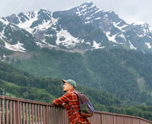 Fototapeta Young man hiker in a red plaid shirt and cap stands by a railing, gazing at majestic snow-capped mountains, surrounded by lush green forests, capturing a moment of tranquility in nature