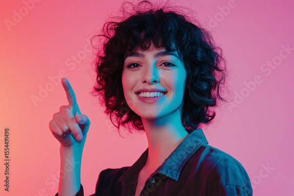Fototapeta Woman with curly hair smiling and pointing upward against a pink and blue gradient background.