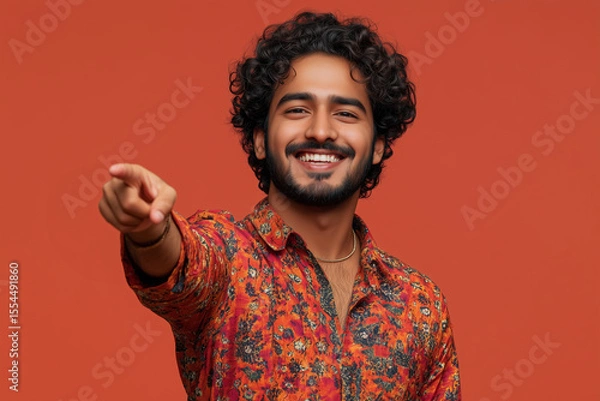 Fototapeta A smiling man with curly hair and a beard, wearing a colorful patterned shirt, points directly at the camera against a solid orange background.