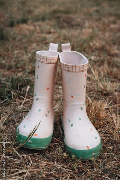 Obraz  A pair of small white children's wellington boots with green soles and playful prints, slightly dirty from soil, standing on dry grass in an outdoor setting