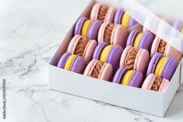 Fototapeta Close up of multicolored macaroons in the cardboard box on the white marble background. Assortment of pink and violet macaroon with yellow chocolate filling. Famous French dessert in a gift box