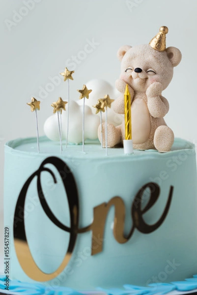 Fototapeta Birthday cake for a little one year old baby with blue chocolate frosting decorated with chocolate figure of teddy bear and white clouds. Baby shower cake for a little boy on turquoise background