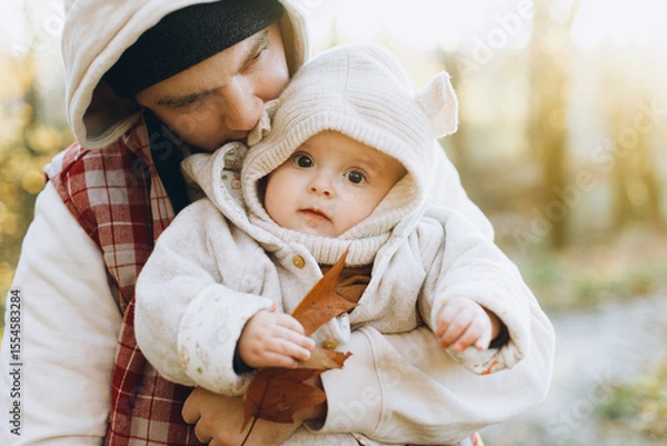 Obraz Happy father hugging and kissing cute baby son in soft evening light in autumn woods. Fatherhood. Stylish man with adorable child in cozy warm sweaters embracing in fall park