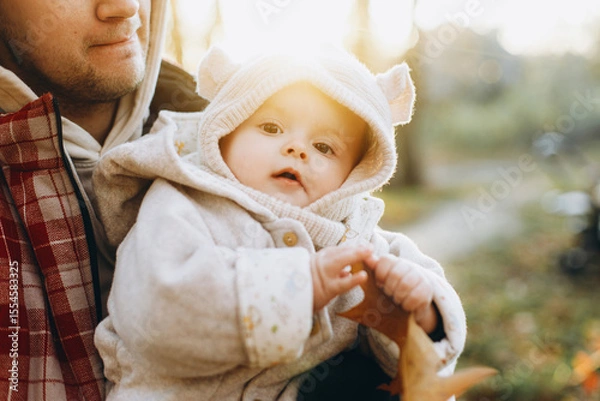 Obraz Happy cute baby son holding autumn leaf in fathers hands in soft evening light in autumn woods. Fatherhood. Stylish man with adorable child in cozy warm sweaters hugging in fall park