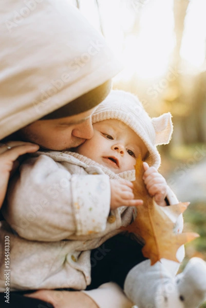 Obraz Happy father hugging and kissing cute baby son in soft evening light in autumn woods. Fatherhood. Stylish man with adorable child in cozy warm sweaters embracing in fall park