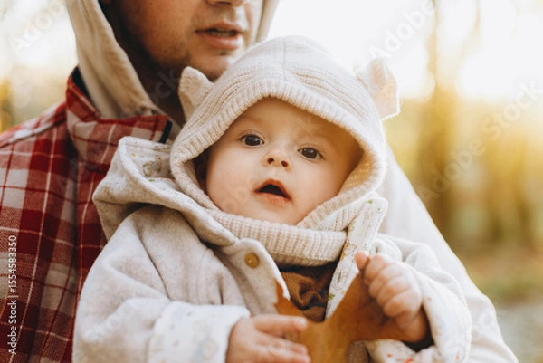 Obraz Happy cute baby son holding autumn leaf in fathers hands in soft evening light in autumn woods. Fatherhood. Stylish man with adorable child in cozy warm sweaters hugging in fall park