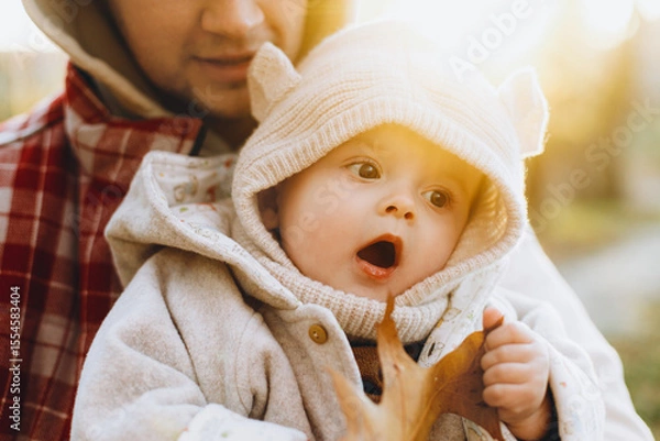 Obraz Happy cute baby son holding autumn leaf in fathers hands in soft evening light in autumn woods. Fatherhood. Stylish man with adorable child in cozy warm sweaters hugging in fall park