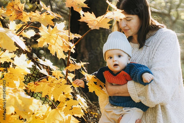 Obraz Happy mother hugging cute baby son in autumn woods with yellow leaves. Motherhood. Stylish woman with adorable child in cozy warm sweaters relaxing in fall park