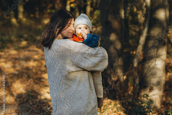 Obraz Happy mother hugging cute baby son in autumn woods. Motherhood. Stylish woman with adorable child in cozy warm sweaters embracing and relaxing in fall park