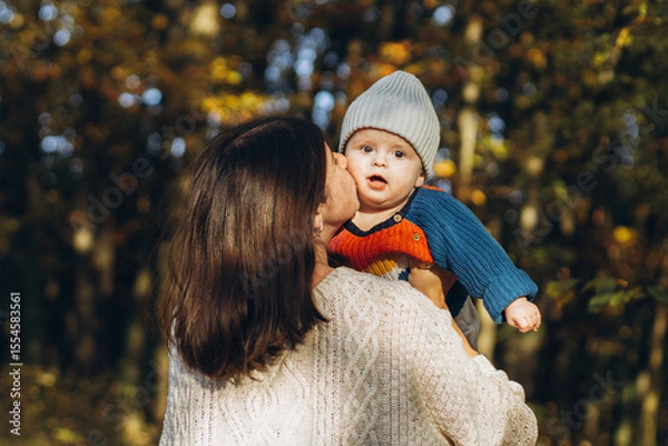 Obraz Happy mother hugging and kissing cute baby son in autumn woods. Motherhood. Stylish woman with adorable child in cozy warm sweaters embracing and relaxing in fall park