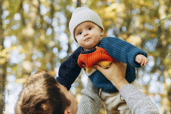 Obraz Cute baby son with funny emotions in father hands close up in autumn woods. Fatherhood. Stylish man with adorable child in cozy warm sweaters having fun and relaxing in fall park
