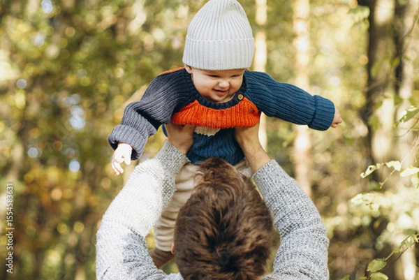 Obraz Cute baby son with funny emotions in father hands close up in autumn woods. Fatherhood. Stylish man with adorable child in cozy warm sweaters having fun and relaxing in fall park