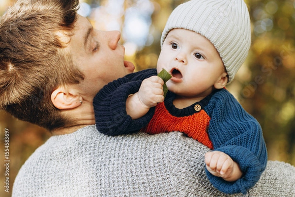 Obraz Cute baby son with funny emotions holding leaves close up in father hands  in autumn woods. Fatherhood. Stylish man with adorable child in cozy warm sweaters relaxing in fall park