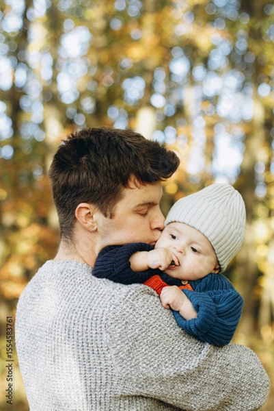 Obraz Happy father hugging and kissing cute baby son in autumn woods. Fatherhood. Stylish man with adorable child in cozy warm sweaters embracing and relaxing in fall park