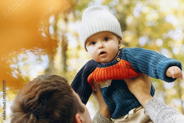 Obraz Cute baby son with funny emotions in father hands close up in autumn woods. Fatherhood. Stylish man with adorable child in cozy warm sweaters having fun and relaxing in fall park