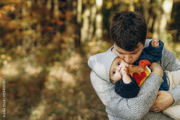 Obraz Happy father hugging and kissing cute baby son in autumn woods. Fatherhood. Stylish man with adorable child in cozy warm sweaters embracing and relaxing in fall park