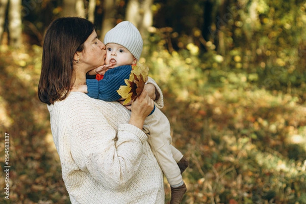 Obraz Happy mother hugging and kissing cute baby son in autumn woods. Motherhood. Stylish woman with adorable child in cozy warm sweaters relaxing in fall park