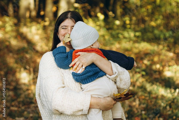 Obraz Happy mother hugging cute baby son in autumn woods. Motherhood. Stylish woman with adorable child in cozy warm sweaters embracing and relaxing in fall park