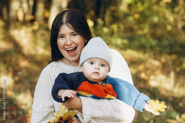Obraz Happy mother hugging cute baby son in autumn woods. Motherhood. Stylish woman with adorable child in cozy warm sweaters embracing and relaxing in fall park