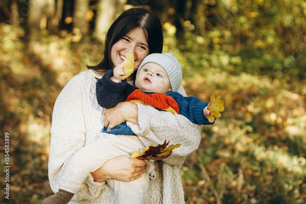 Obraz Happy mother hugging cute baby son in autumn woods. Motherhood. Stylish woman with adorable child in cozy warm sweaters embracing and relaxing in fall park